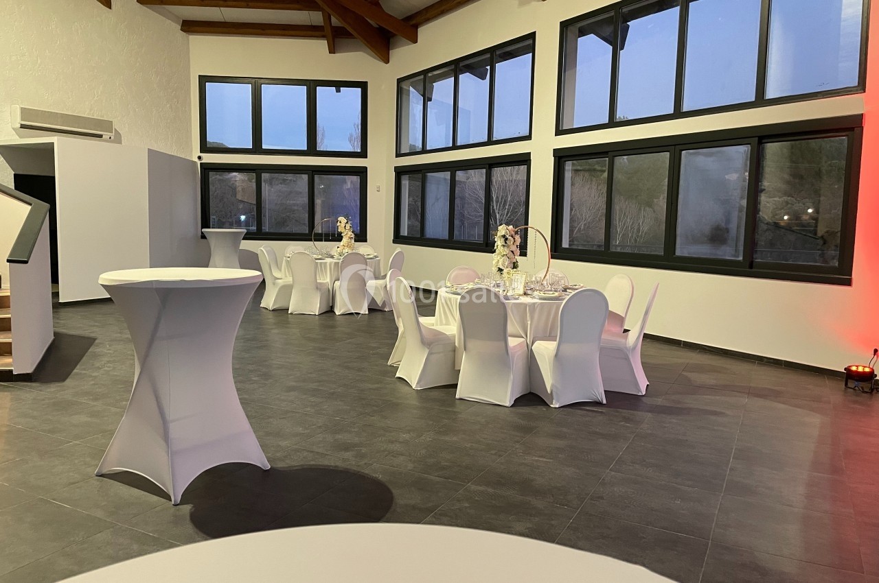 Salle lumineuse avec grandes baies vitrées, tables rondes dressées et chaises blanches, décorée pour un événement.