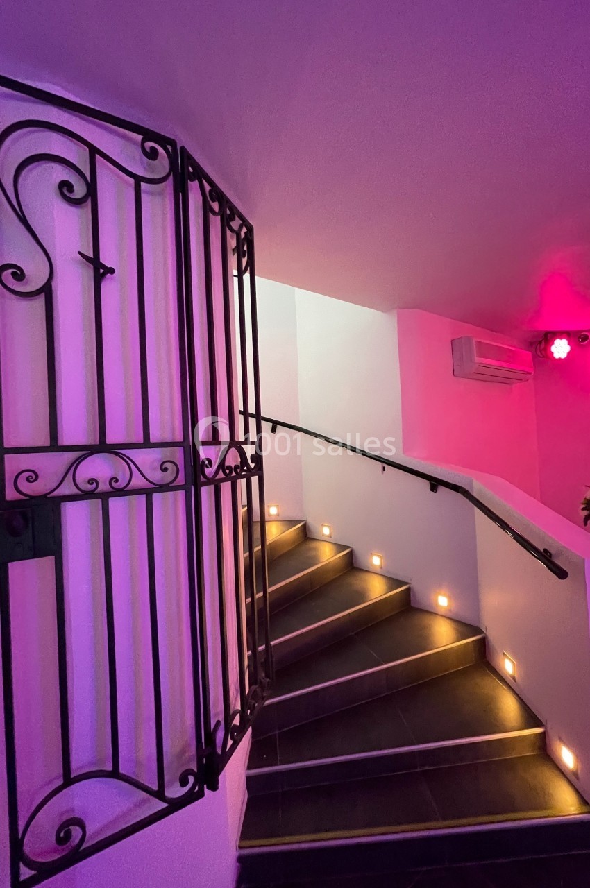 Escalier intérieur éclairé par des lumières au sol, avec une rampe et une porte en fer forgé décorative.