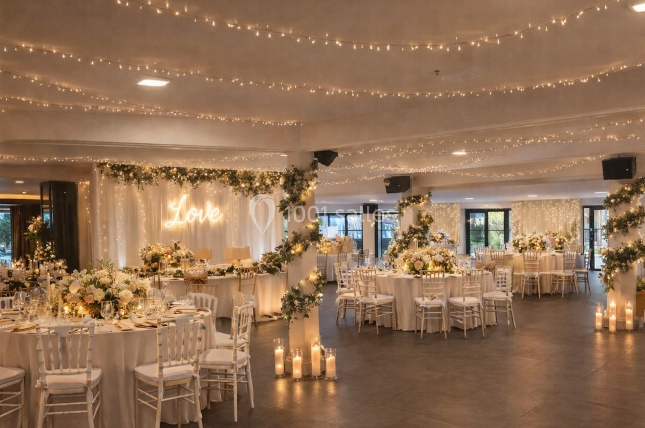 Salle de réception décorée pour un mariage avec guirlandes lumineuses, tables dressées et arrangements floraux élégants.