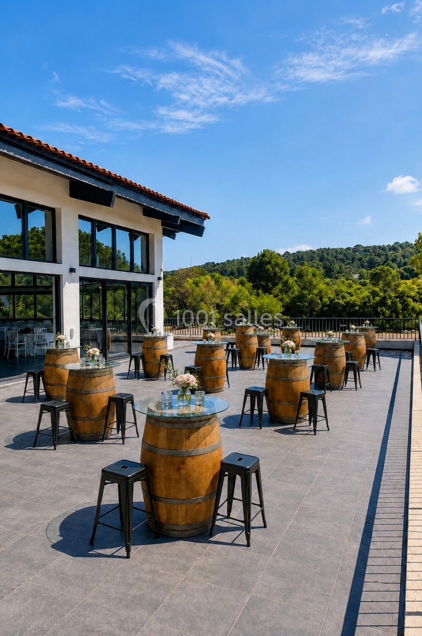 Terrasse ensoleillée avec tables en tonneaux, tabourets noirs et vue sur un paysage verdoyant.