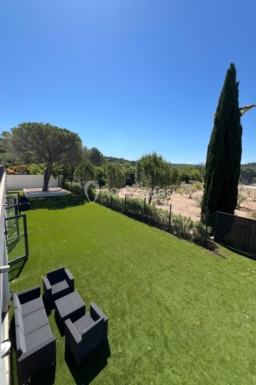 Jardin avec pelouse artificielle, mobilier de jardin noir, clôture et vue sur un paysage arboré sous un ciel dégagé.