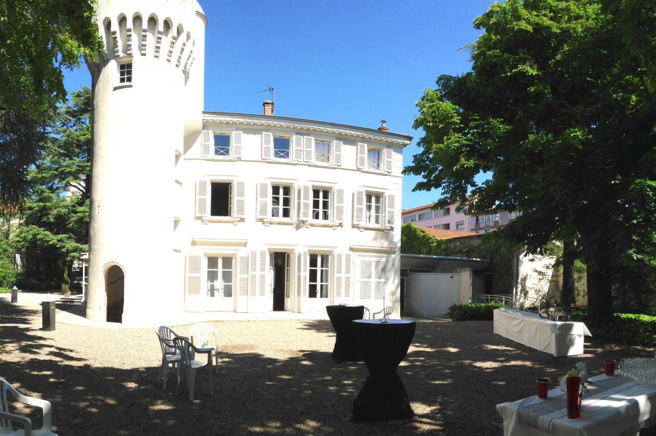 Façade d'une maison blanche avec une tour ronde, entourée d'arbres, tables et chaises disposées dans la cour.