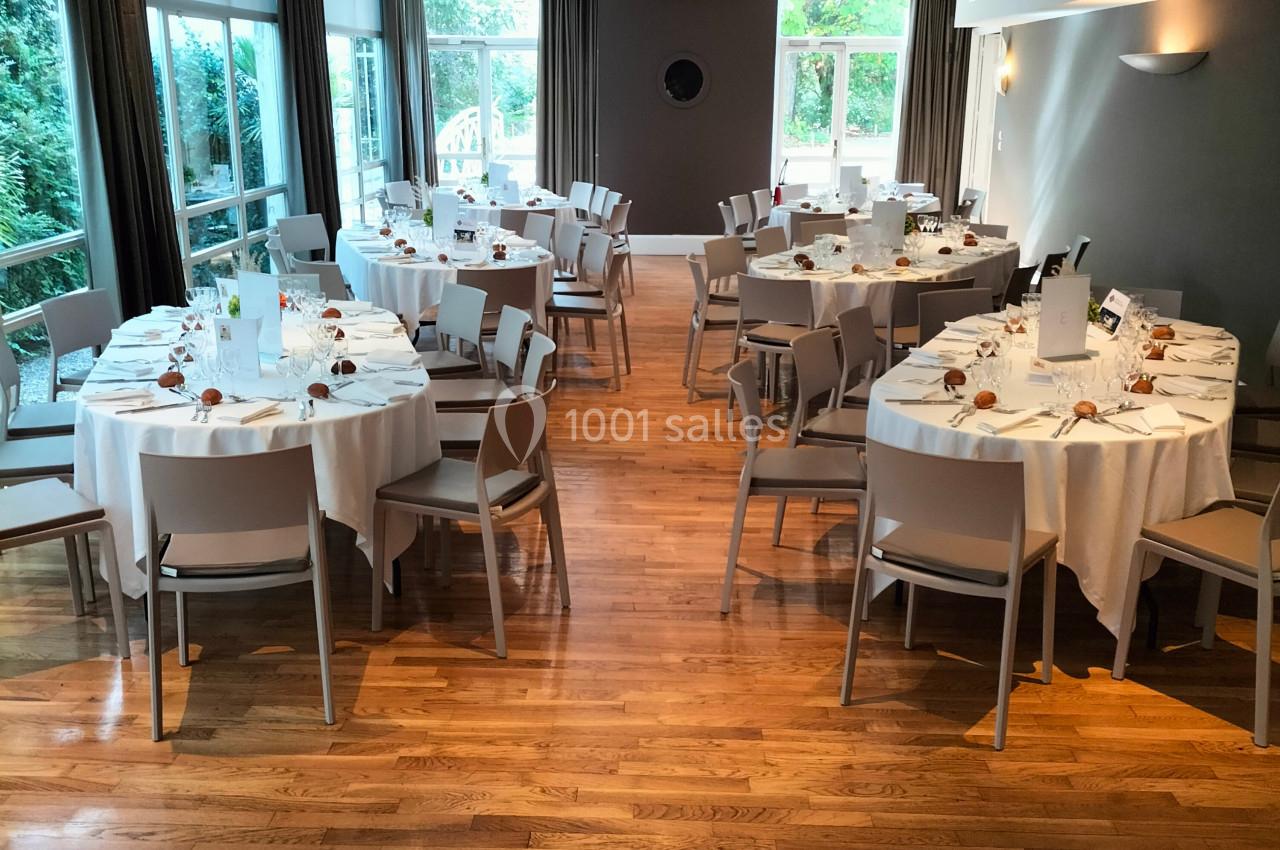 Salle lumineuse avec parquet, tables rondes dressées pour un repas, chaises blanches et grandes fenêtres donnant sur l…