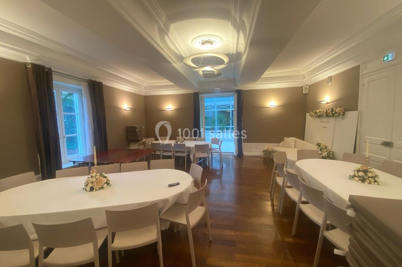 Salle élégante avec tables rondes et chaises blanches, décorée de bouquets, éclairée par des luminaires modernes.