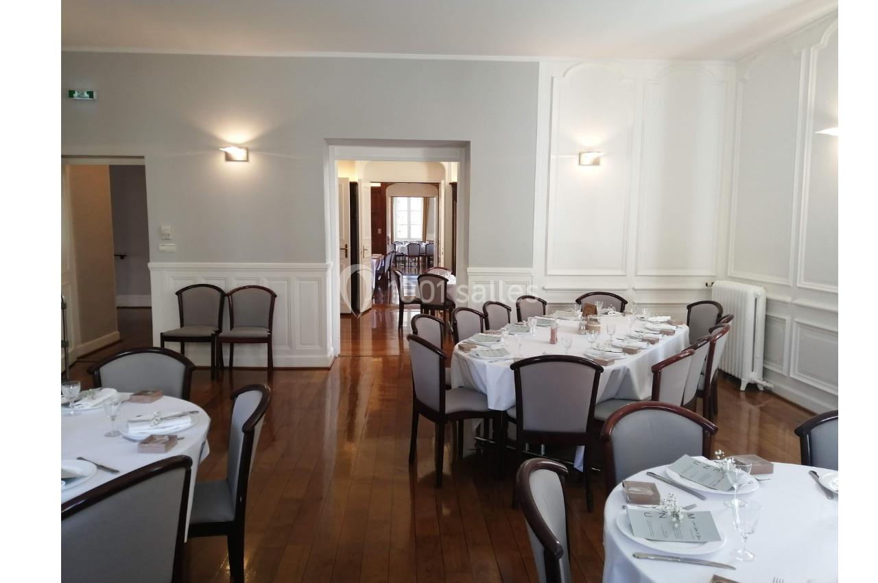 Salle de réception lumineuse avec tables rondes dressées, chaises en bois et murs blancs ornés de moulures.