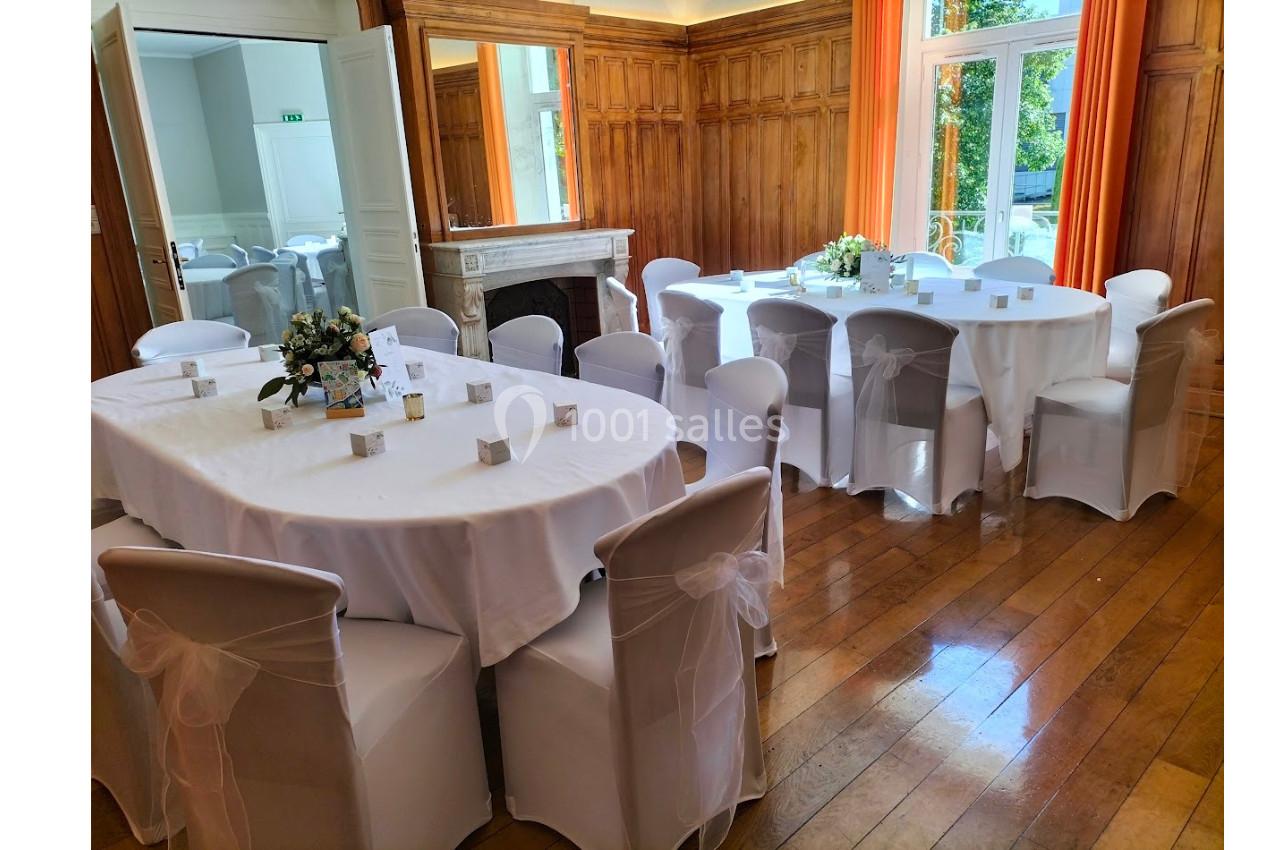 Salle élégante avec tables rondes et ovales dressées de nappes blanches, chaises décorées et lumière naturelle.