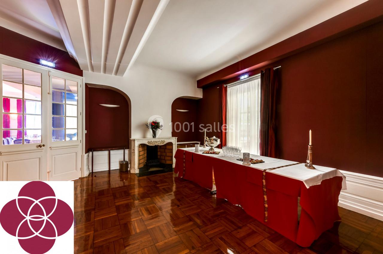 Salle élégante avec parquet, murs rouges, cheminée, table dressée avec nappes blanches et grandes fenêtres.
