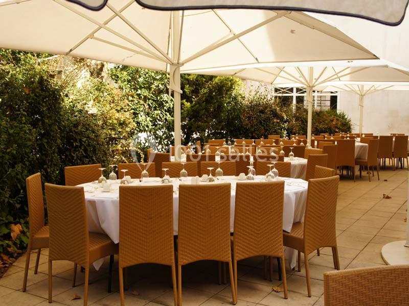 Terrasse aménagée avec des tables rondes dressées, entourées de chaises en osier, sous des parasols blancs.