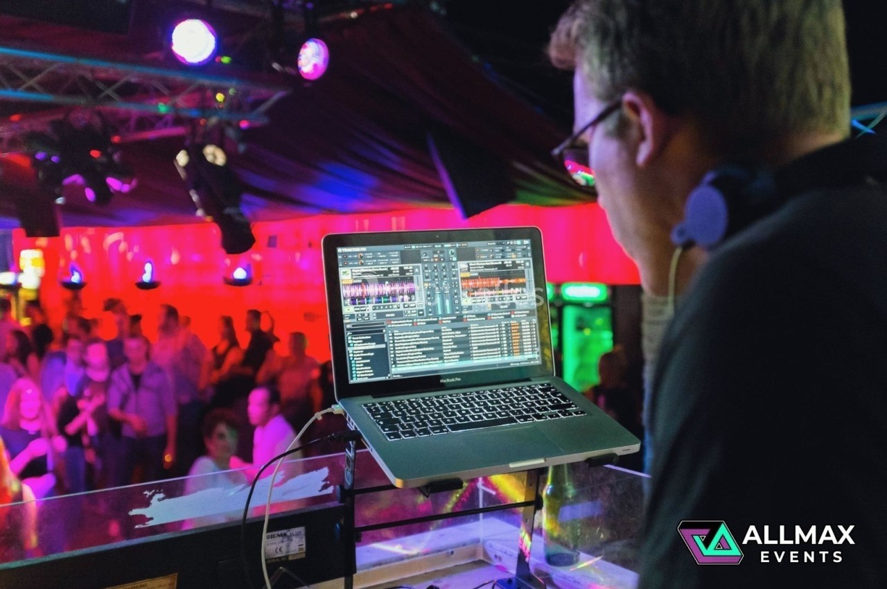 Un DJ mixe de la musique sur un ordinateur portable dans une salle éclairée par des lumières colorées, avec un public en…