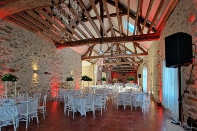 Salle de réception avec poutres apparentes, murs en pierre, tables dressées et éclairage chaleureux.