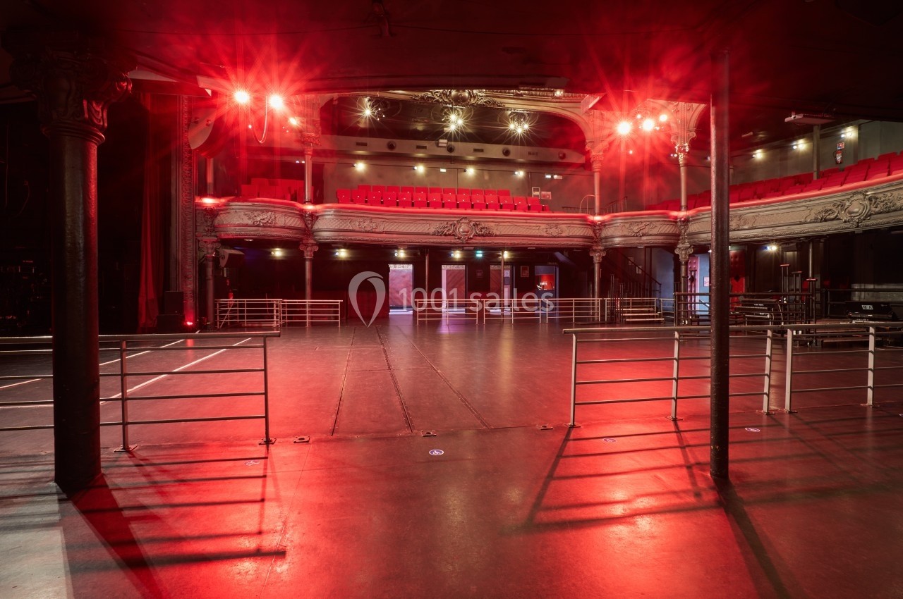 Salle de spectacle vide avec balustrades, scène et balcons ornés, éclairée par des lumières rouges.
