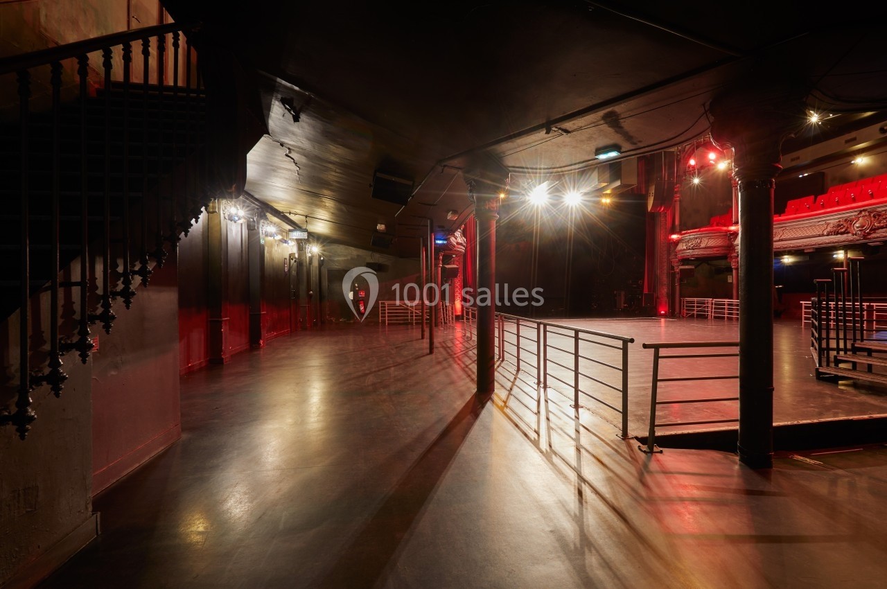 Salle de spectacle vide avec éclairage tamisé, balustrades métalliques et escalier en colimaçon à gauche.