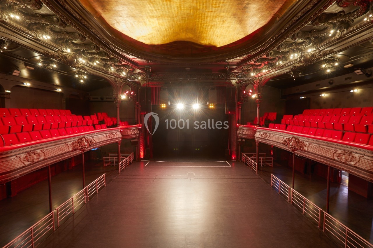 Salle de théâtre vide avec balcons ornés, sièges rouges et scène éclairée par des projecteurs.