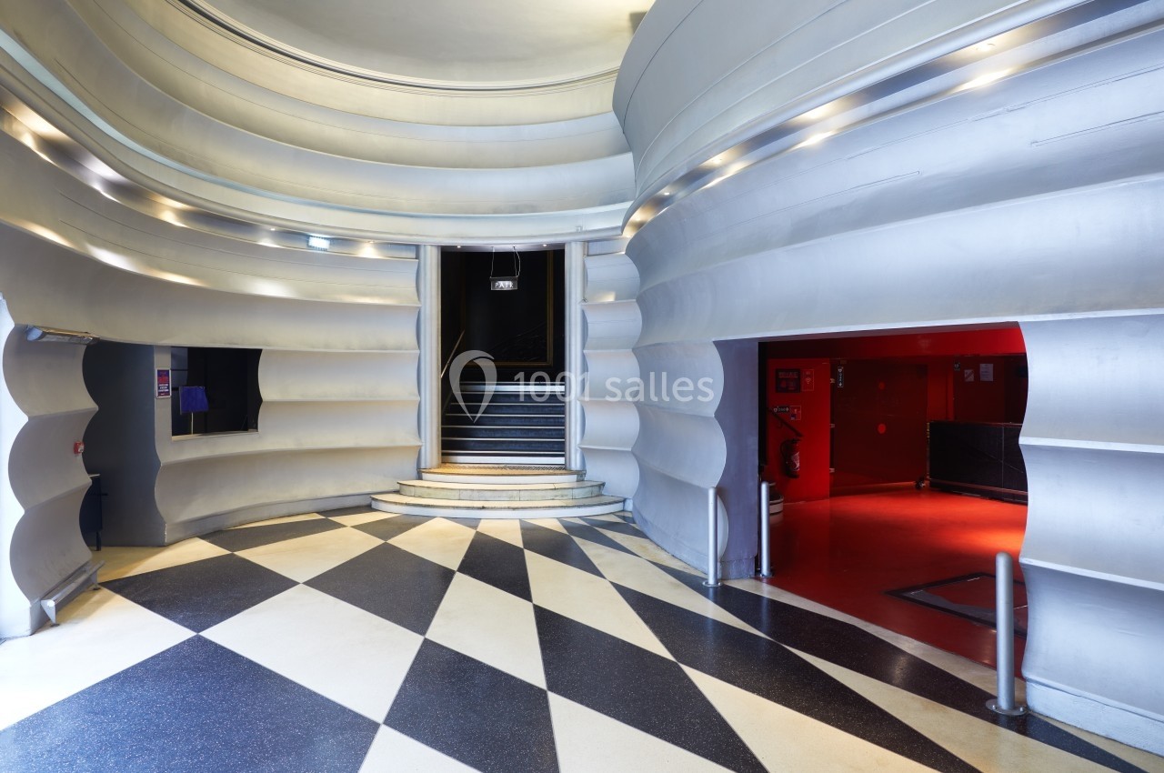 Hall d'entrée avec murs aux formes ondulées, sol à motifs géométriques et escalier menant à une porte noire.