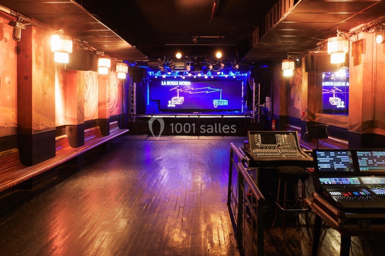 Salle de concert vide avec scène éclairée, équipements audio en premier plan et murs décorés.