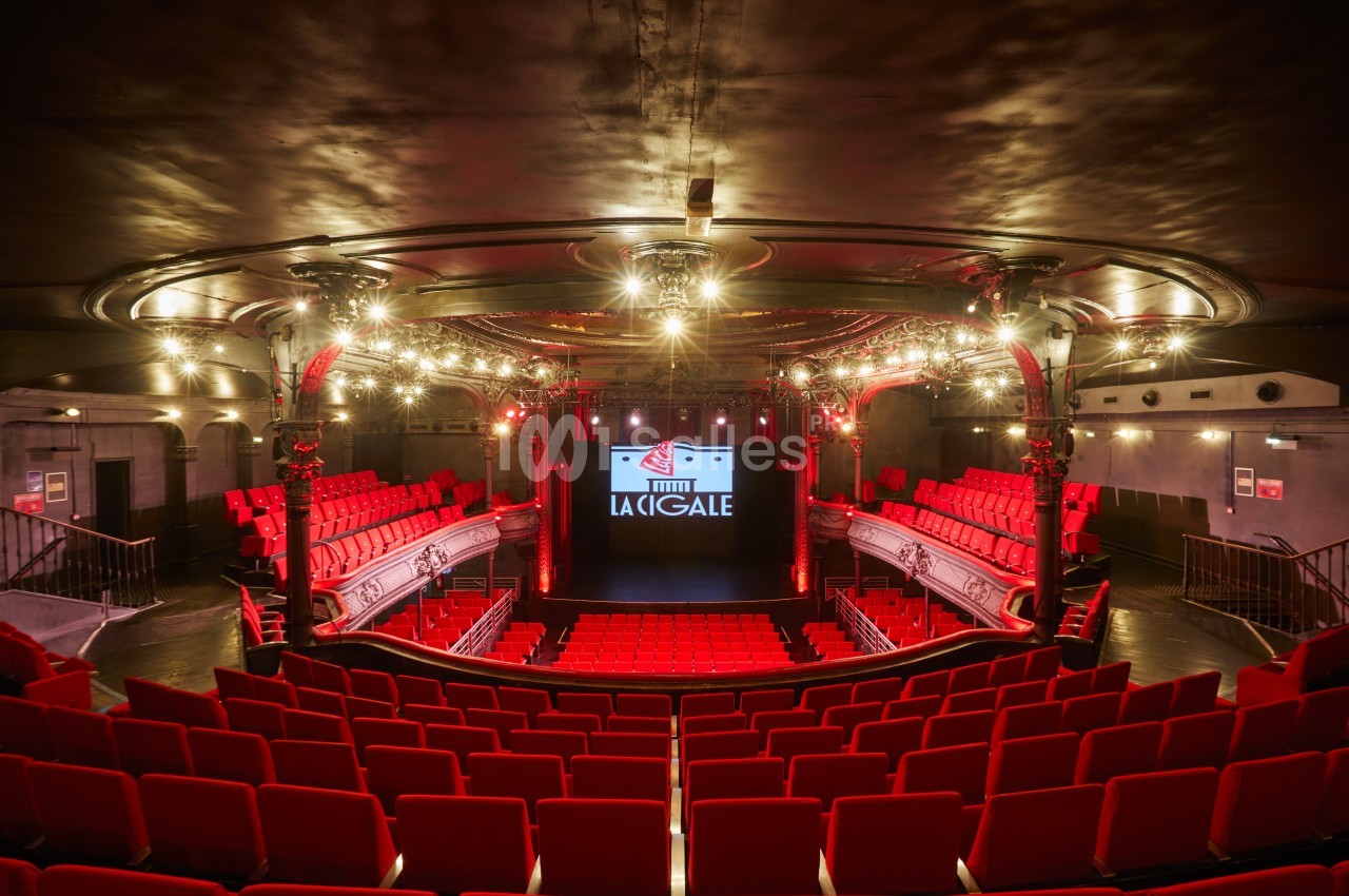 Salle de spectacle avec sièges rouges, balcon, scène éclairée et enseigne ’La Cigale’ visible en arrière-plan.