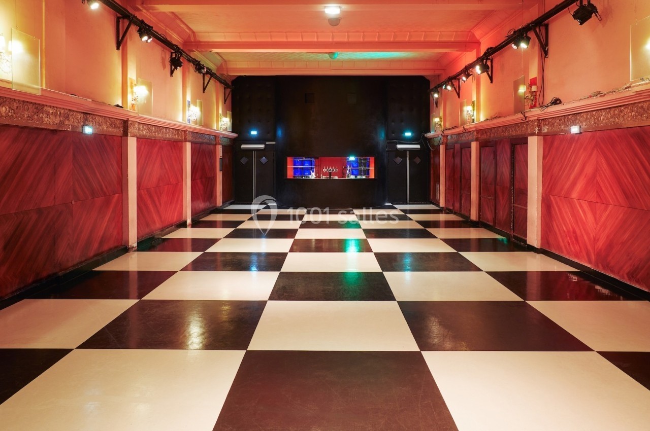 Salle vide avec sol à damier noir et blanc, murs rouges ornés de moulures et bar éclairé en arrière-plan.