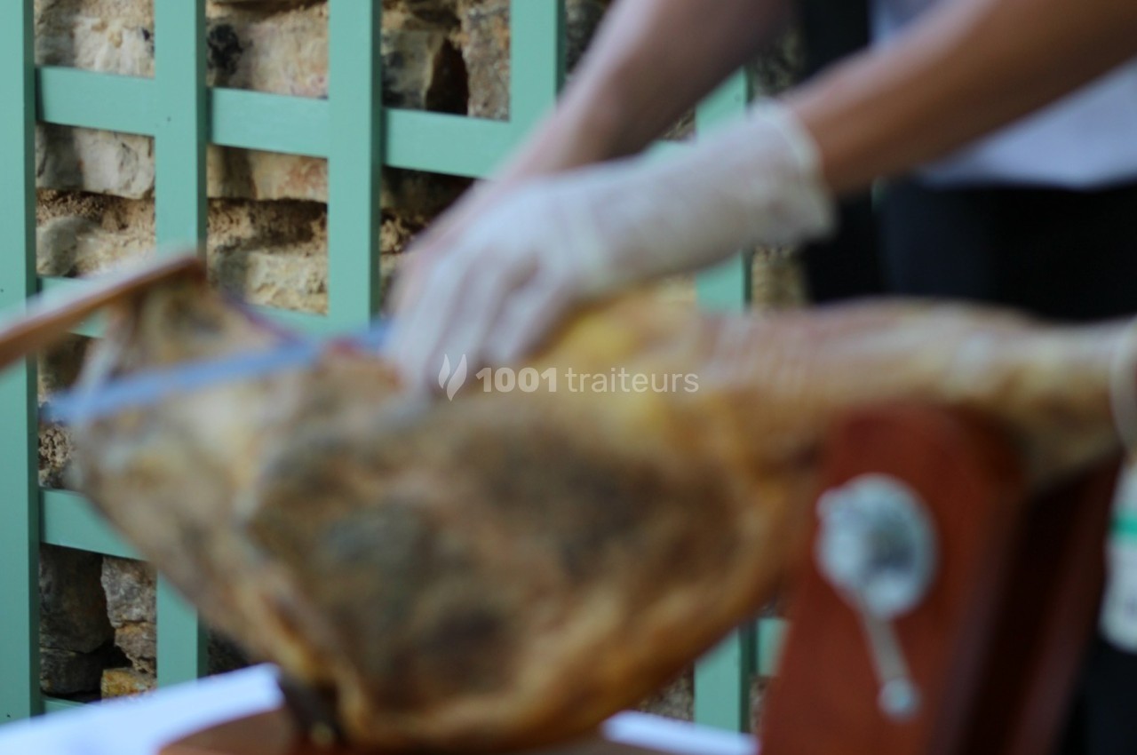 Personne découpant un jambon sur un support en bois, portant des gants, avec une barrière en arrière-plan.