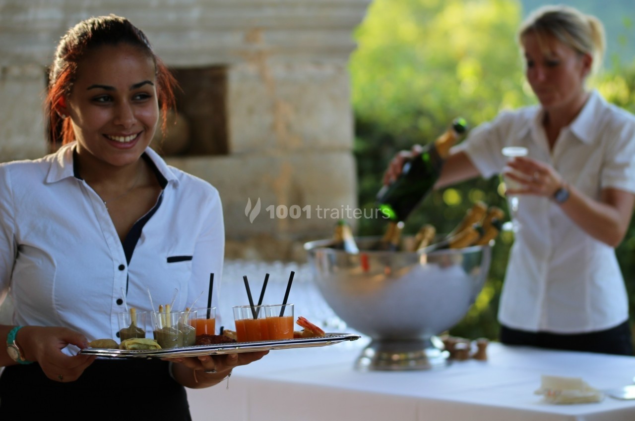 Serveuse souriante portant un plateau de boissons, avec une collègue servant du champagne en arrière-plan.