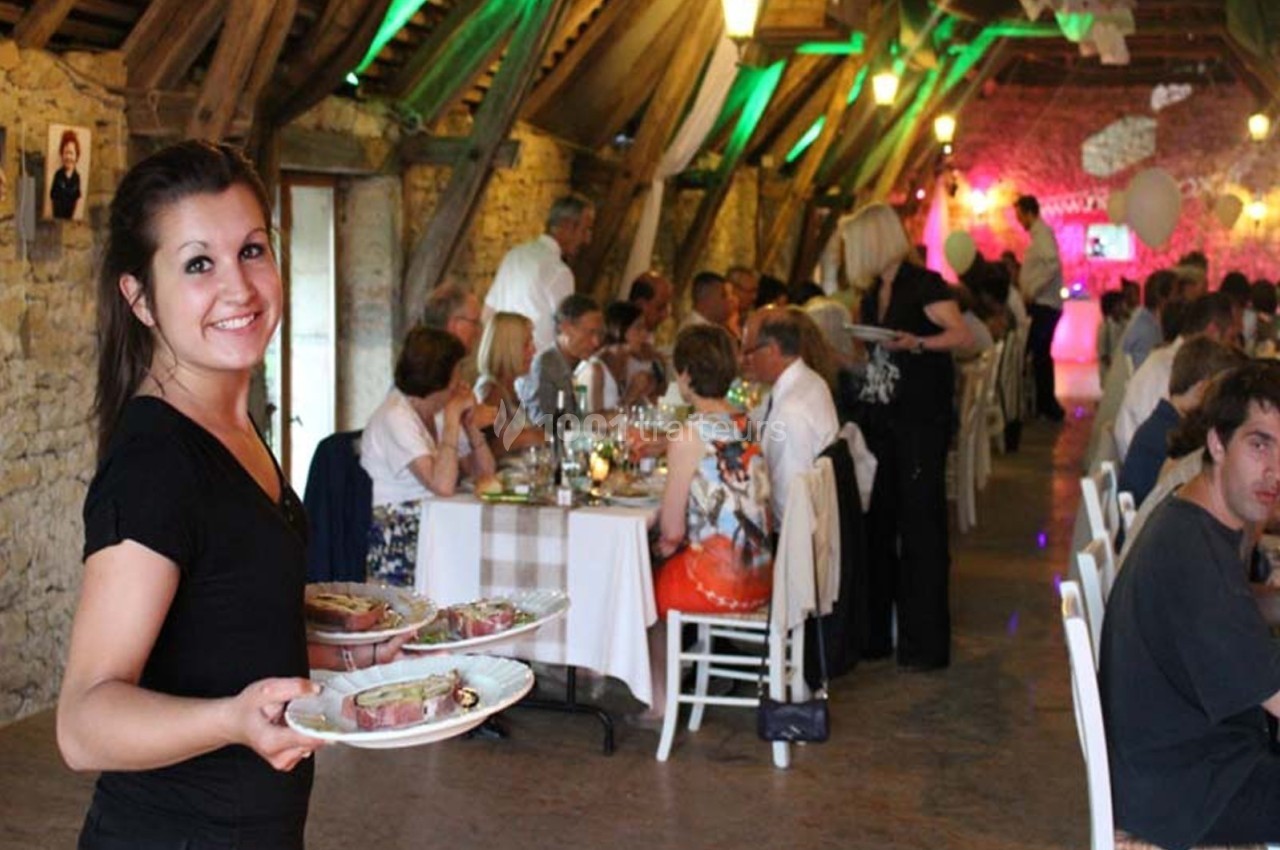 Serveuse souriante apportant des plats dans une salle de réception rustique avec des invités attablés.