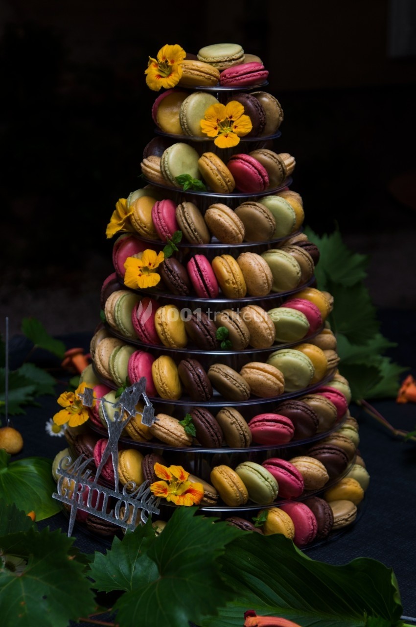 Tour de macarons colorés décorée de fleurs jaunes, présentée sur une table sombre avec des feuilles en arrière-plan.