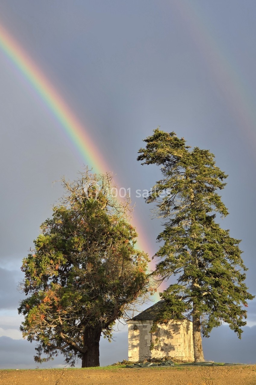 Un petit bâtiment en pierre entouré de deux arbres, avec un arc-en-ciel visible dans un ciel nuageux.