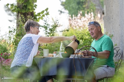 Salon lumineux avec murs en pierre, cheminée, canapé, table ronde avec chaises jaunes, et vue sur une cuisine ouverte.