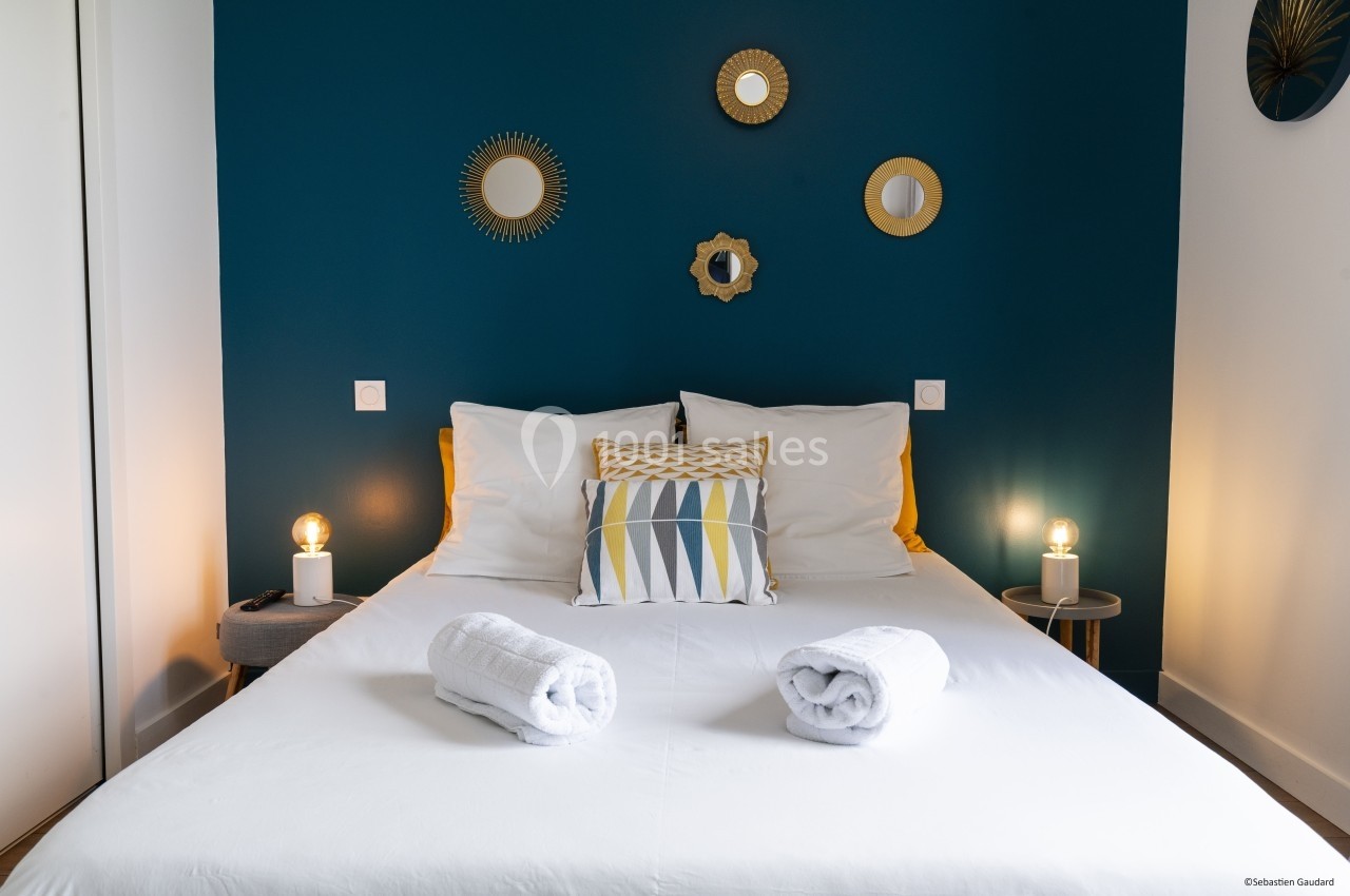 Lit double avec coussins et serviettes pliées, encadré par deux lampes de chevet, devant un mur bleu décoré de miroirs.