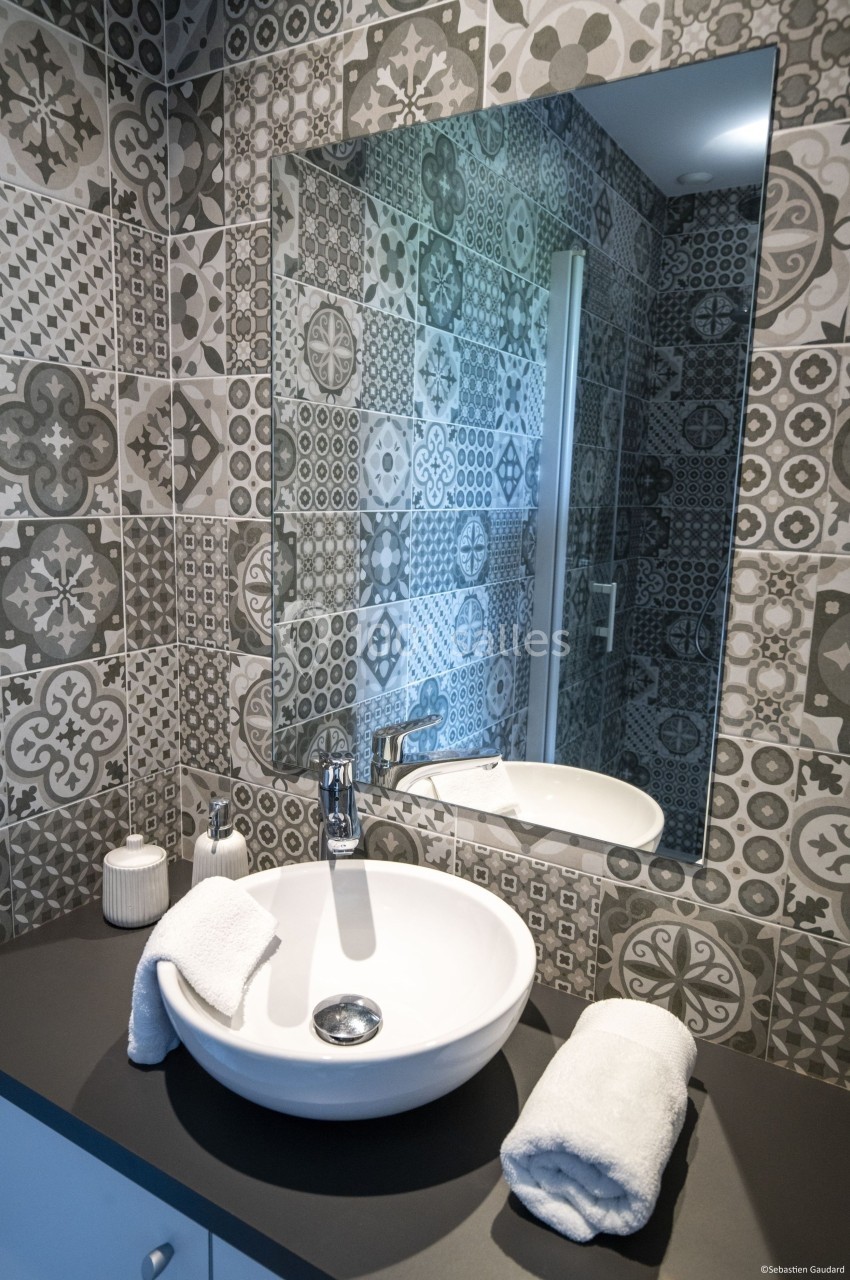 Lavabo blanc avec miroir, posé sur un plan de travail sombre, entouré de carreaux de faïence à motifs géométriques.