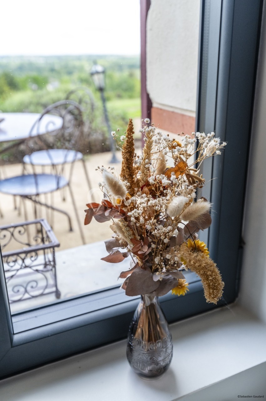 Bouquet de fleurs séchées dans un vase en verre posé sur un rebord de fenêtre avec vue sur une terrasse et un paysage.