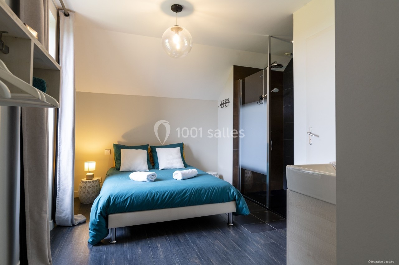 Chambre lumineuse avec lit double, linge de lit bleu, salle de bain attenante et sol en parquet foncé.