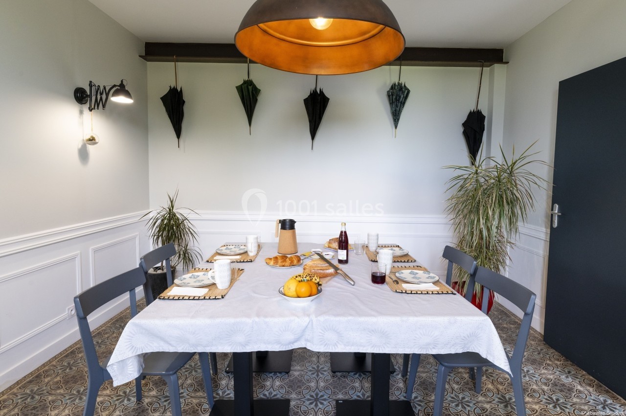 Salle à manger avec une table dressée pour un repas, décorée de plantes, d'un luminaire suspendu et de parapluies accrochés…
