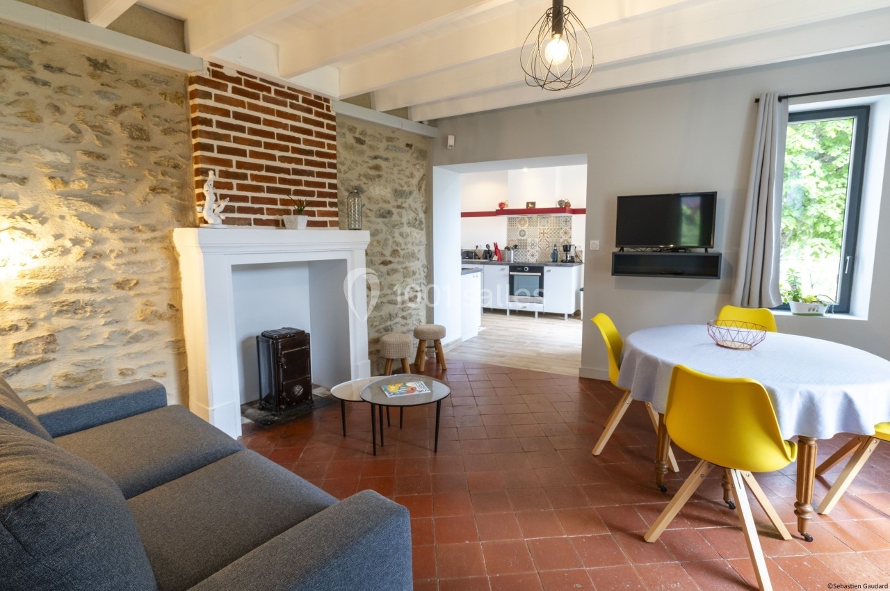 Salon lumineux avec murs en pierre, cheminée, canapé, table ronde avec chaises jaunes, et vue sur une cuisine ouverte.