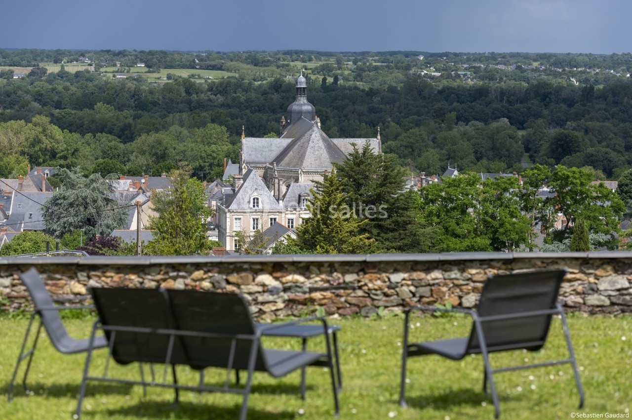 Vue d'un village avec une église au centre, entouré de verdure, depuis un jardin avec des chaises en premier plan.