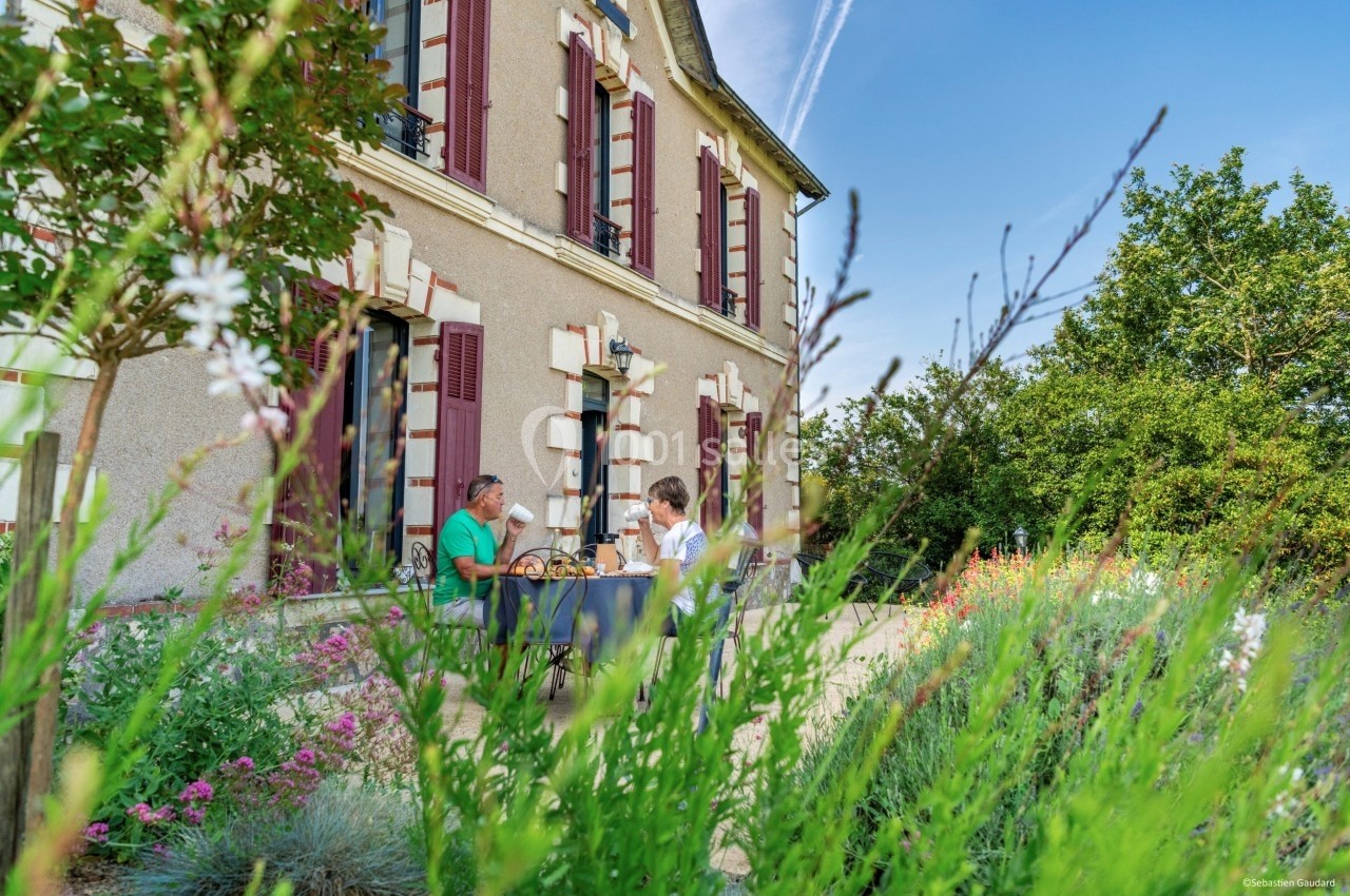 Deux personnes prennent un repas en terrasse devant une maison en pierre, entourée de fleurs et de verdure.
