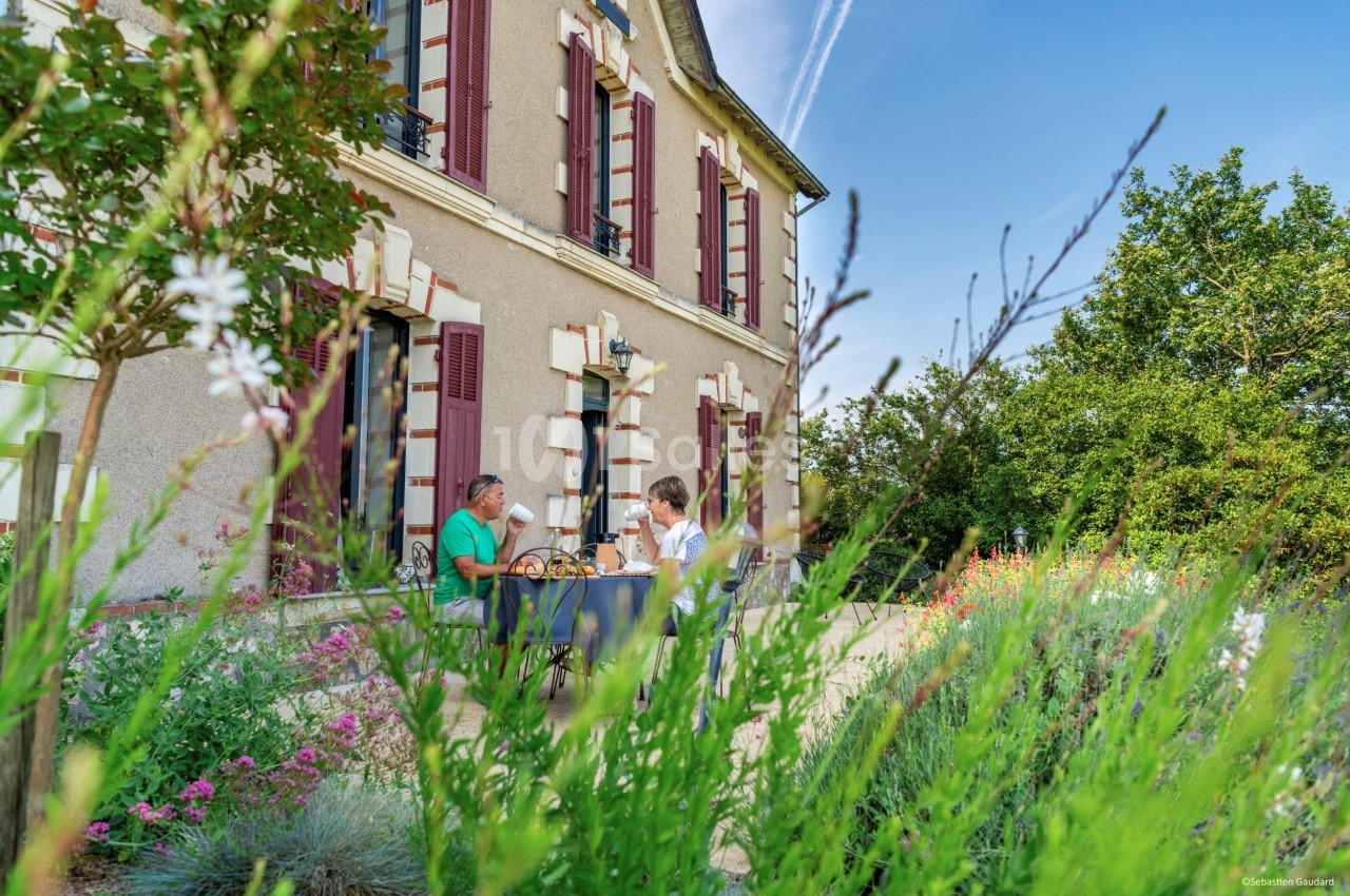 Deux personnes prennent un repas en terrasse devant une maison en pierre, entourée de fleurs et de verdure.