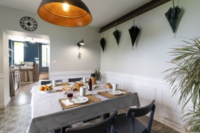 Salon lumineux avec murs en pierre, cheminée, canapé, table ronde avec chaises jaunes et vue sur une cuisine ouverte.