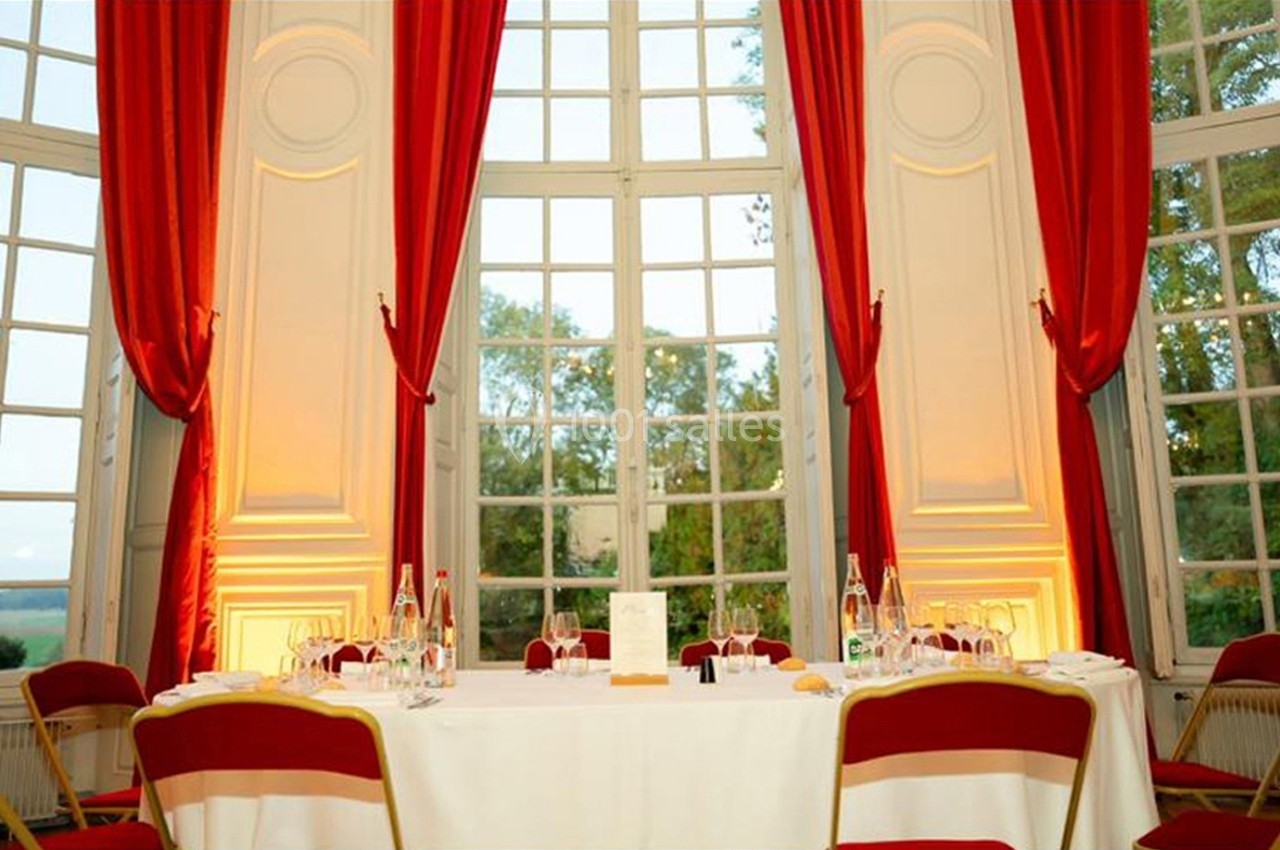 Table dressée avec nappes blanches, chaises rouges et fenêtres hautes encadrées de rideaux rouges dans une salle lumineuse.