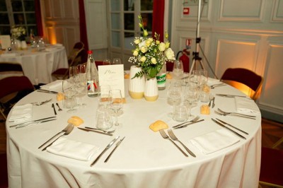 Table ronde dressée pour un repas, avec nappes blanches, couverts, verres, pains et centre de table floral.