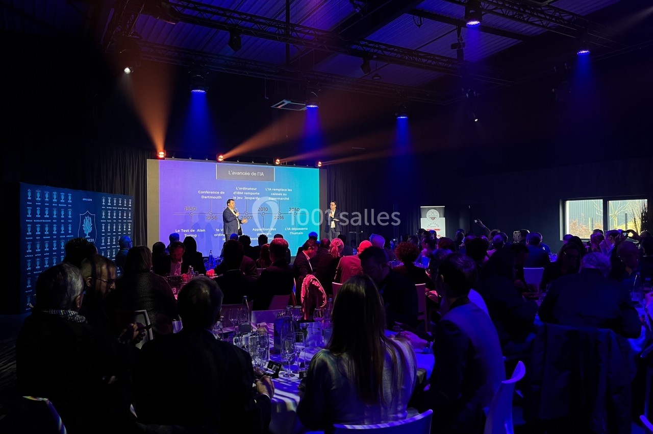 Salle de conférence avec des participants assis, éclairage coloré et deux intervenants sur scène devant un écran.