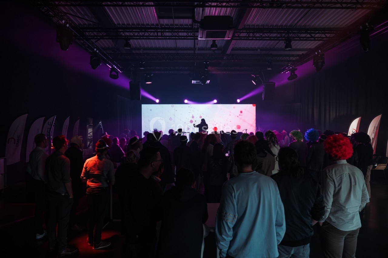 Une foule assiste à une présentation sur scène dans une salle sombre éclairée par des lumières colorées.