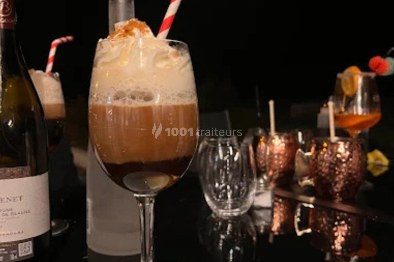 Verre de cocktail avec mousse et paille rouge et blanche, posé sur une table noire avec d'autres verres en arrière-plan.