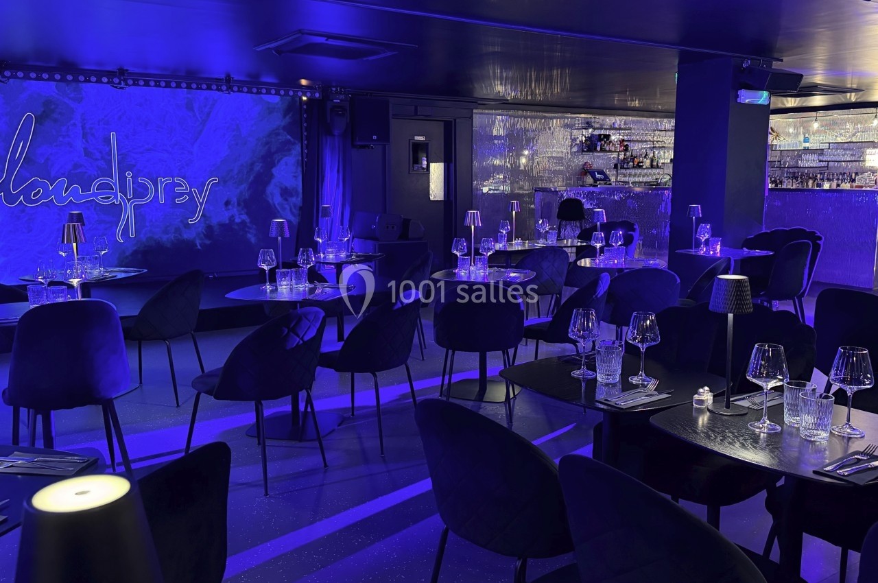 Salle de restaurant moderne avec éclairage bleu, tables dressées et bar illuminé en arrière-plan.