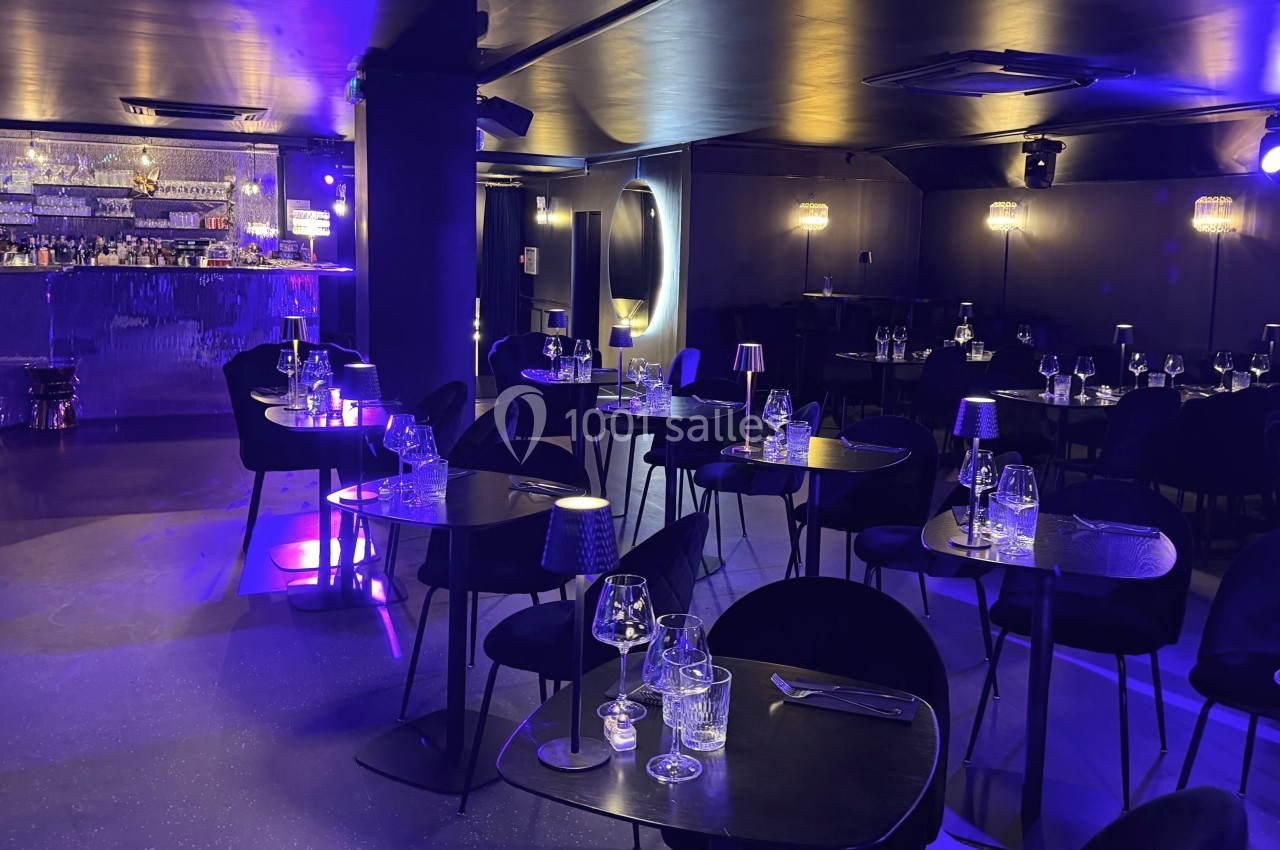 Salle de restaurant moderne avec éclairage tamisé violet, tables dressées et un bar en arrière-plan.