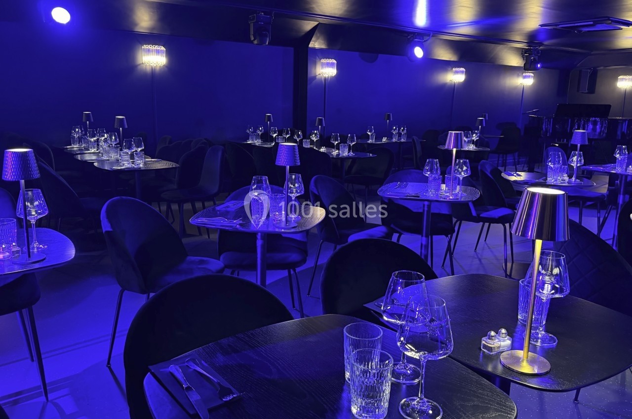 Salle de restaurant élégante avec tables dressées, éclairage tamisé bleu et petites lampes sur chaque table.