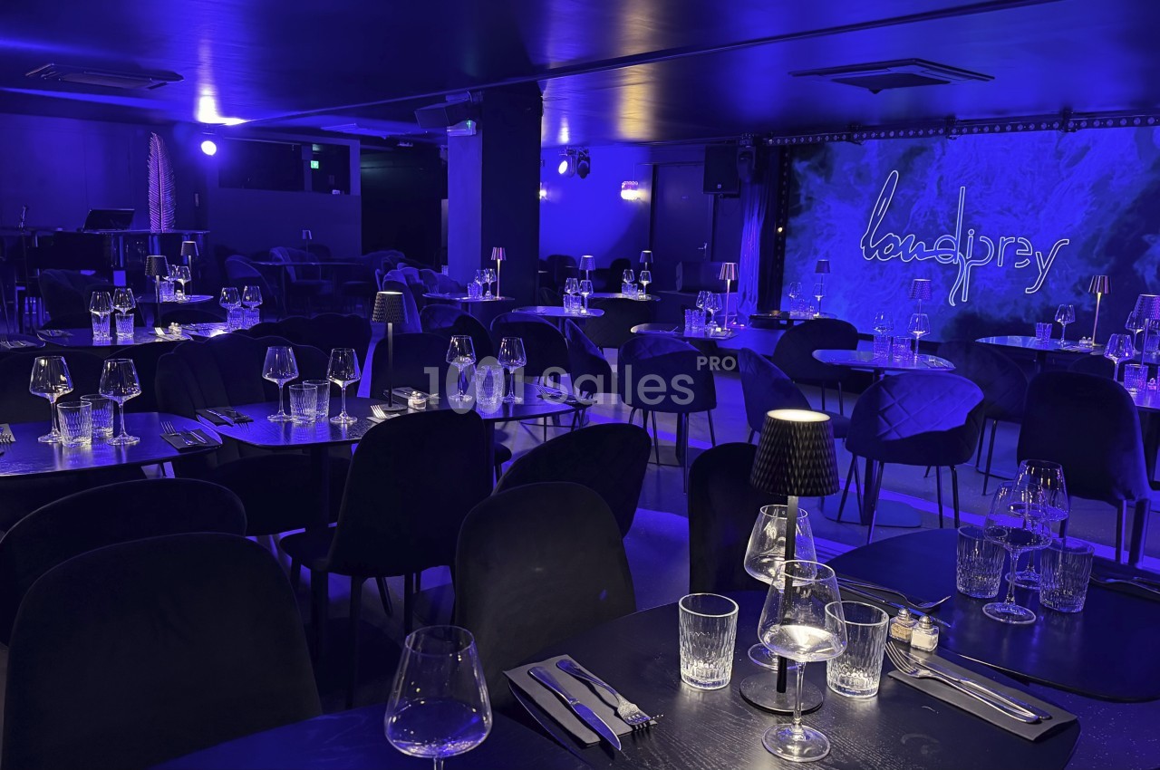 Salle de spectacle avec tables dressées, éclairage tamisé bleu et scène en arrière-plan.