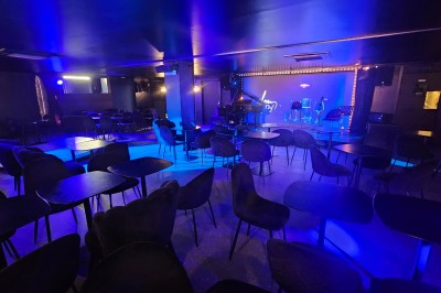 Miniature Location salle Paris 10 (Paris) - Lou Diprey #7 Salle de spectacle avec scène éclairée en bleu, équipée d'un piano, de micros et de chaises devant des tables.