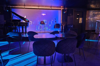 Miniature Location salle Paris 10 (Paris) - Lou Diprey #13 Salle de spectacle avec scène éclairée en bleu, équipée d'un piano, de micros et de chaises devant des tables.