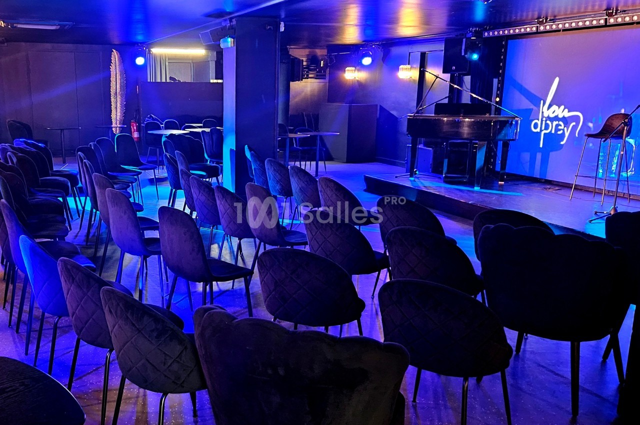 Salle de spectacle avec sièges alignés, éclairage bleu et scène équipée d'un piano à queue et d'un micro.