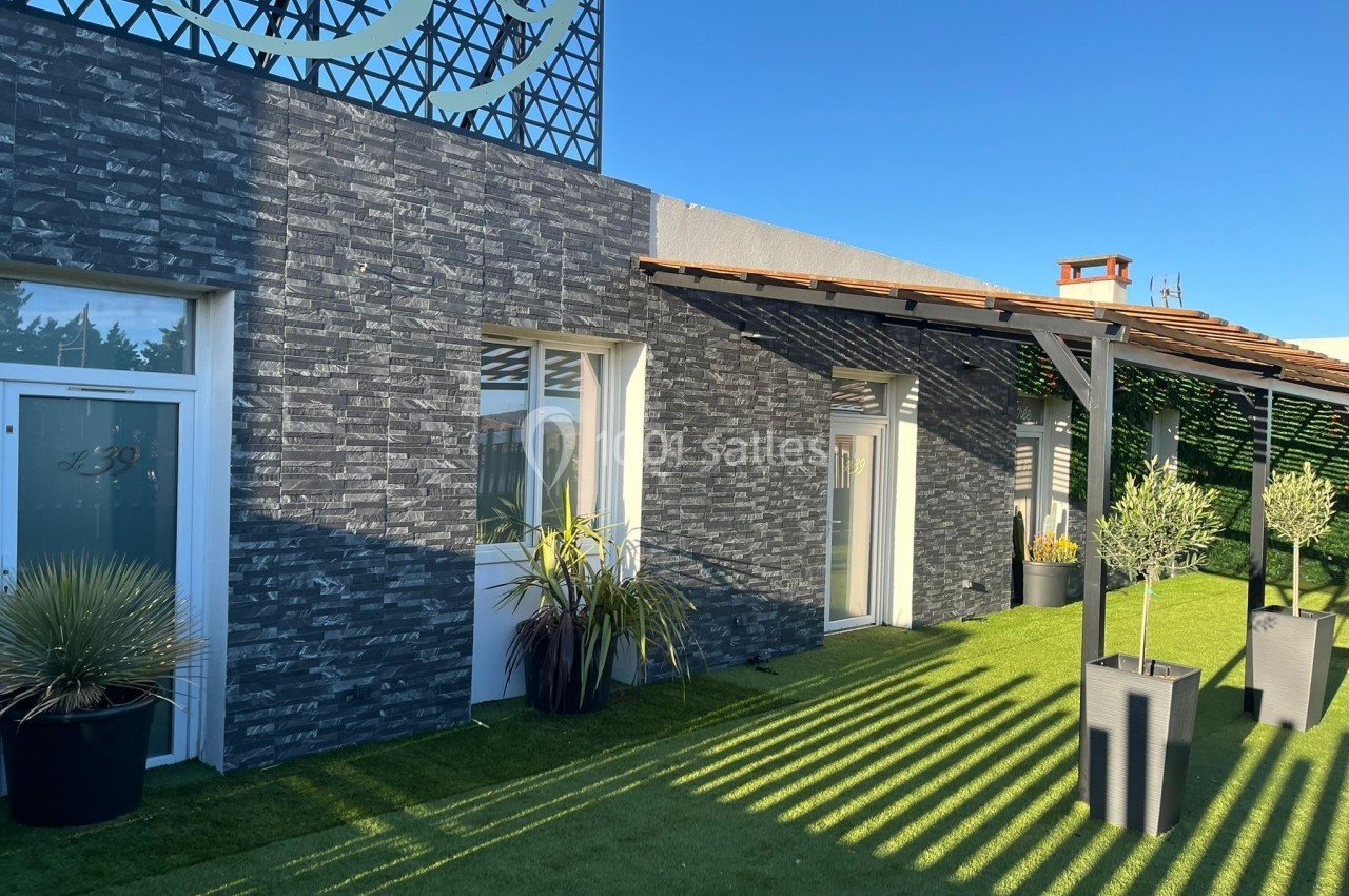 Façade moderne avec murs en pierre grise, portes vitrées et terrasse aménagée avec plantes sur gazon synthétique.